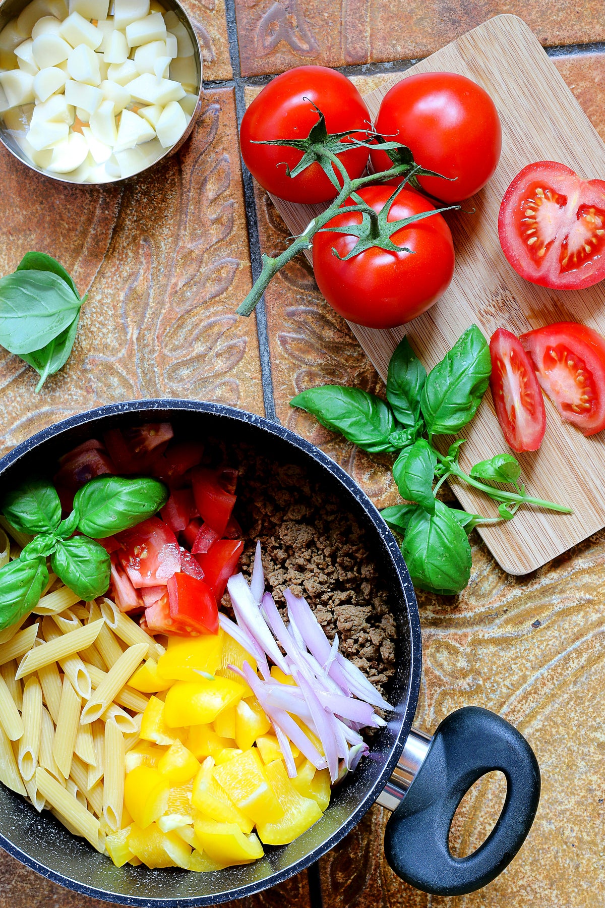 Eine Bowl mit Nudeln und Gemüse in einem Topf und daneben aufgeschnittene Tomaten auf einem Brett dekoriert mit Basilikum